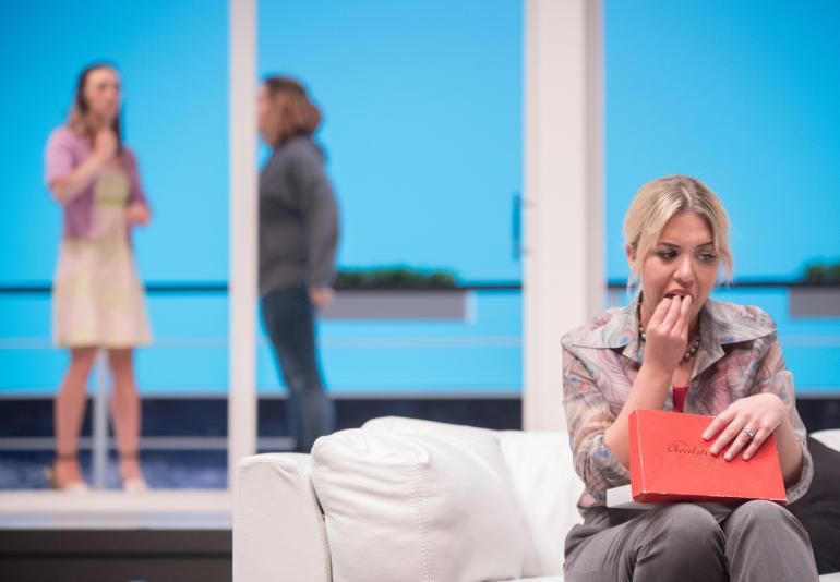 female cast member sits on couch eating chocolates, other cast members can be seen on stage behind her