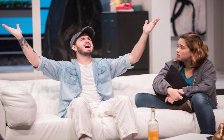 cast member sits on couch with hands raised, other cast member looks at him