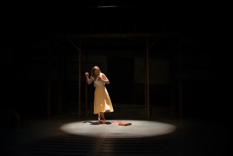 female cast member stands in spotlight on dark stage and looks down at book on the ground