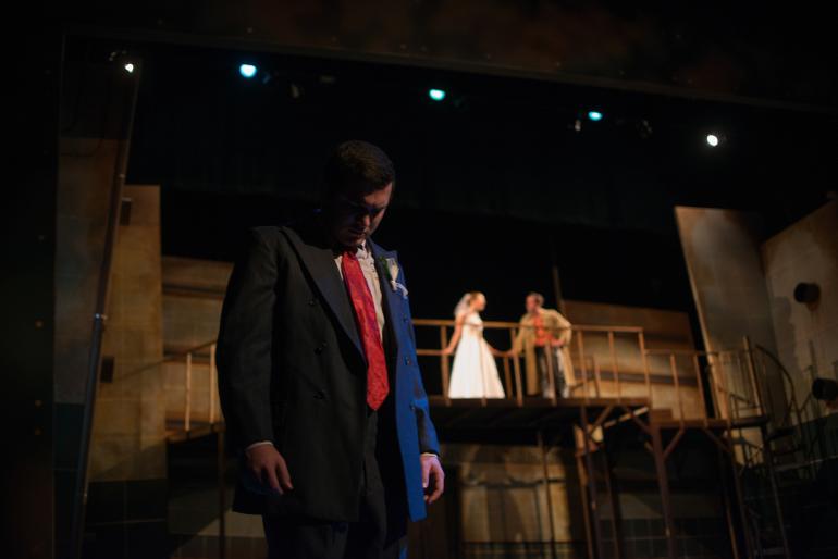 man stands at front of stage with head down, behind him up on a platform a female character in wedding dress talks to male character