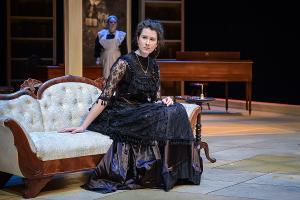 female character sits on couch in black dress, other female character stands at back of stage behind her