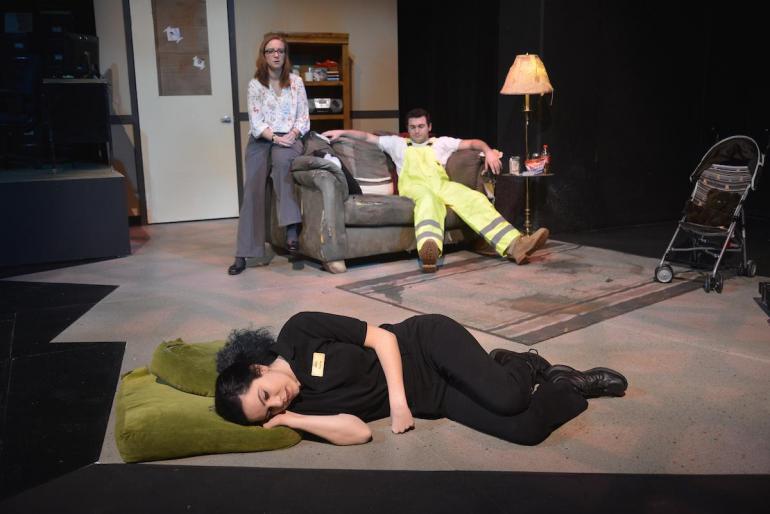 female cast member lays on floor, other cast members sit on couch behind her