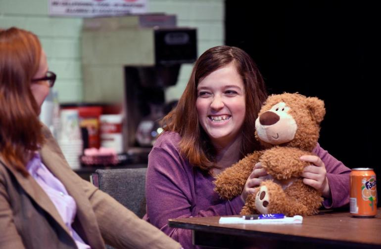 two female cast members sit at a table, one holds a teddy bear and smiles
