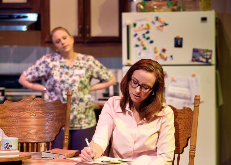 woman sits a table writing, other female cast member stands behind her in a kitchen setting