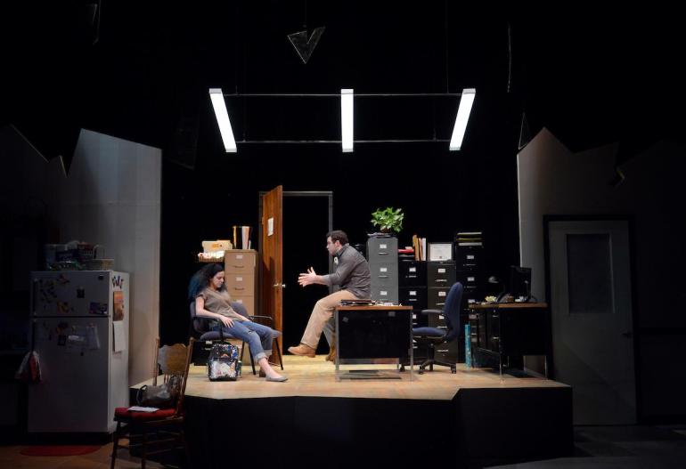 female sits in chair and male sits on desk on stage set that looks like an office