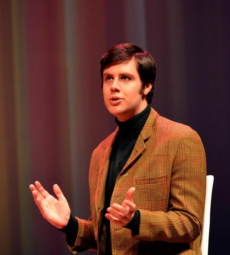 male character dressed in blazer and black turtle neck talks/sings