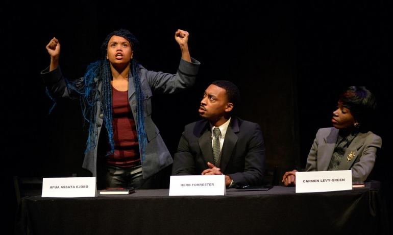 Cast member stands on stage with her arms up, with other cast members sitting at judges table looking at her