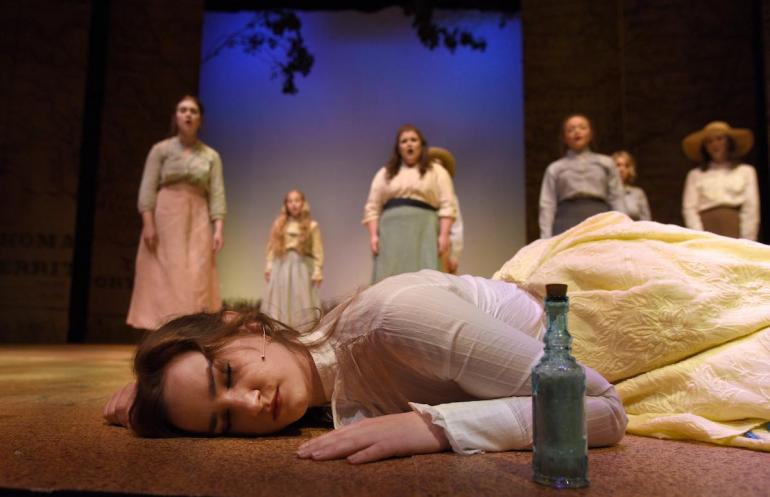 close up of female cast member laying on stage with bottle on ground in front of her, other female cast members stand behind her singing