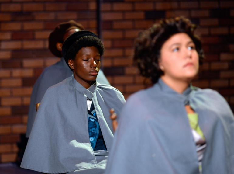 female cast members in matching coats sit on stage