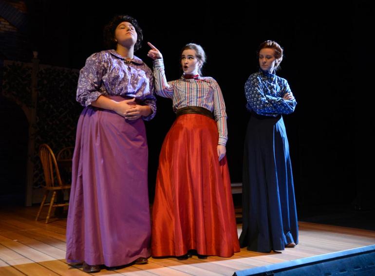 three female cast members stand on stage in dresses, one points into distance