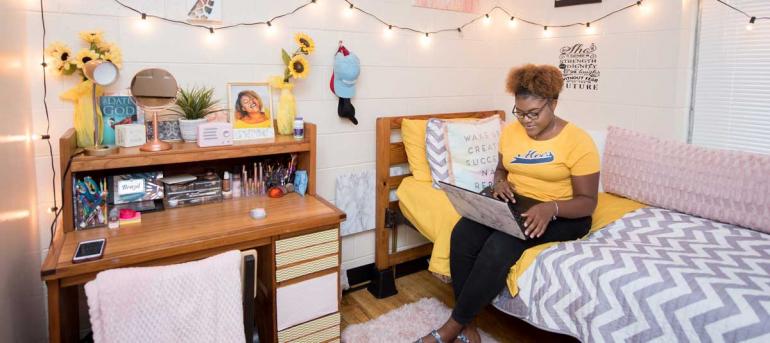 Girl sits in bed with laptop in her bedroom at boling apartments.