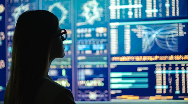 Silhoutted student wearing glasses looks at wall of screens with data graphs and charts.