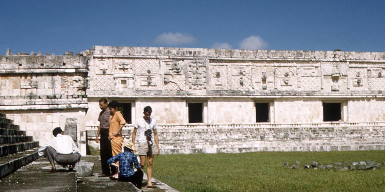 Five people grouped on the steps of an ancient Latin American archaeological site with a stone edifice featuring ornate decoration in the background.