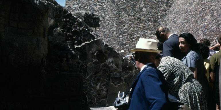 A group of people at the base of an ancient archaeological stone structure with an animal motif. A person holds a vintage camera at the front of the group.