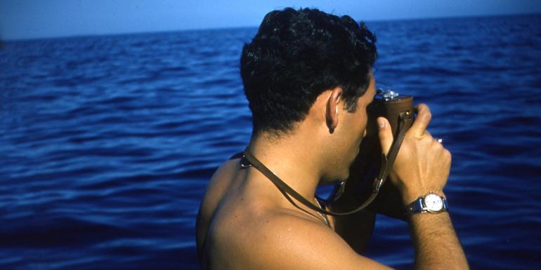 The back of a man, looking out towards the a vibrant blue sea, and holding a camera to his face with the camera strap around his neck. 