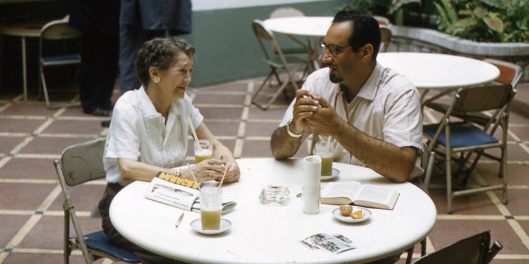 A man and woman sit at a round table talking to each other. The table and chairs are aluminum, and there is a glass filled with green liquid on the table.