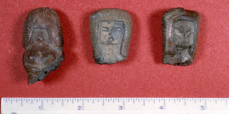 Three figurine heads laid out on a solid red background. The figurines each have different facial features, but they are all of similar size and material. The faces are carved into an earth-tone clay using line to depict facial structure. 