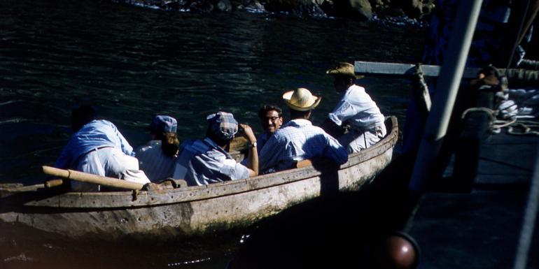 Six people wearing hats and light-colored collared shirts seated in a canoe in the water.