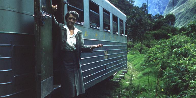 Woman wearing a dress suit stands on the foot rail of a train peering out to the lush forest and mountains of South America. 