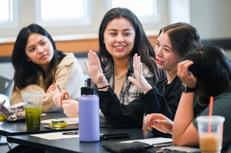 Four young women talk in a college classroom.