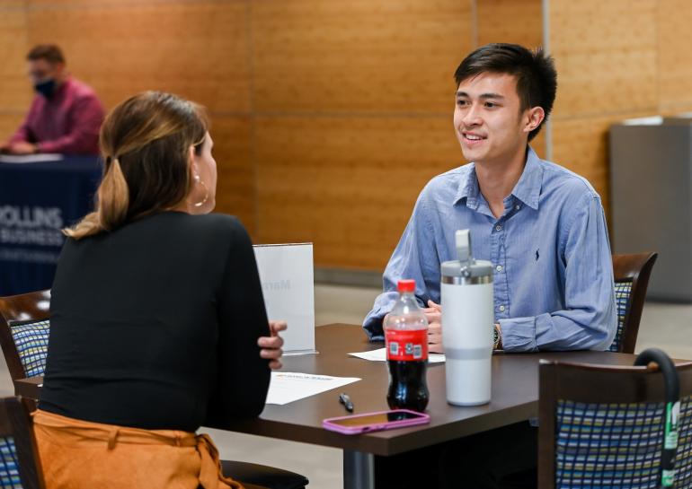Our 100% online Master of Science in Management provides leadership skills for professional success. Student in a blue button down shirt talking with a woman in a black jacket.