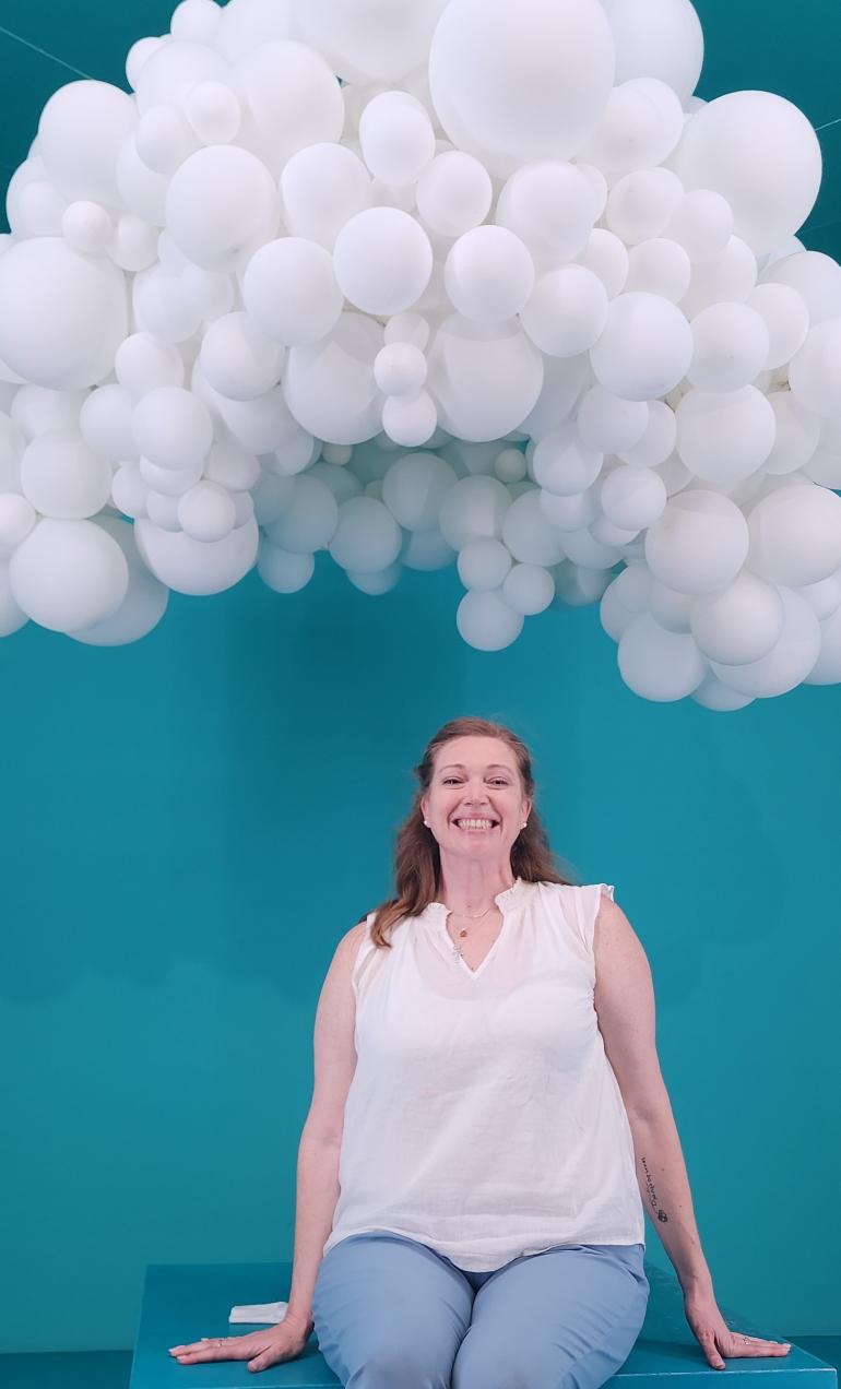 Jamie Newberg Jamie Newberg smiling while standing against a blue wall, wearing a white dress and standing under white balloons.