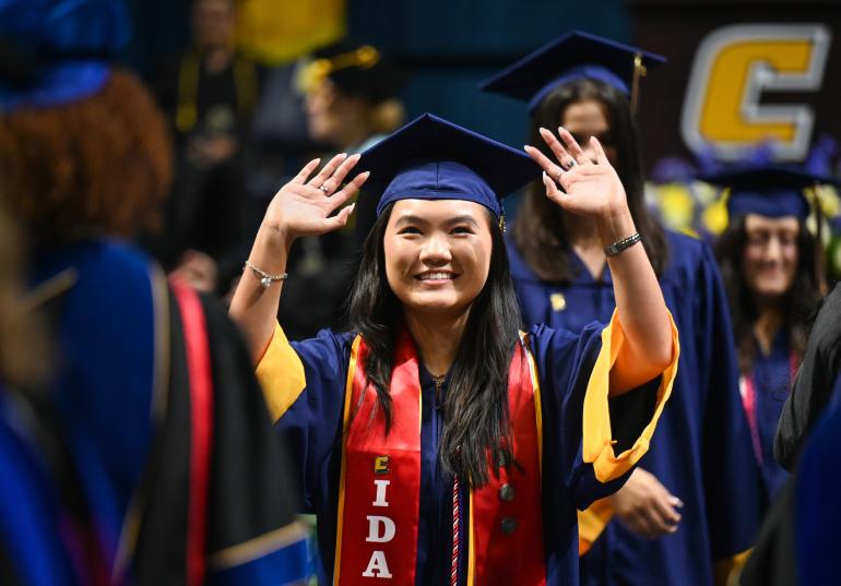 UTC graduate student waring a cap and gown with her hands raised in the air.