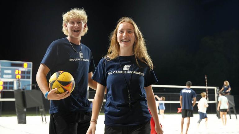 Picture of 2 students at the sand volleyball courts.  One is holding a volleyball.