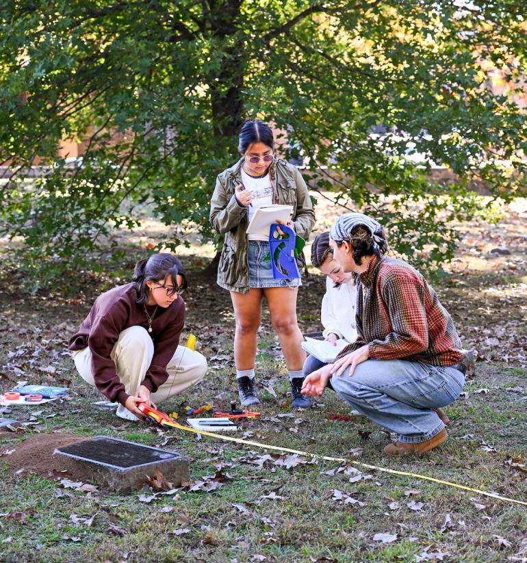 Three UTC students conduct an outdoor environmental science lab, measuring and recording data on a grassy area with fallen leaves.