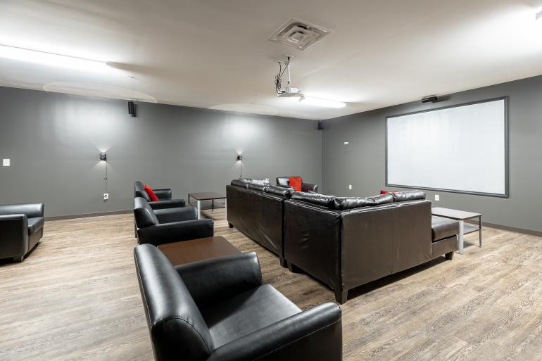 Gray room with large white screen on wall black couches and chairs through out the space