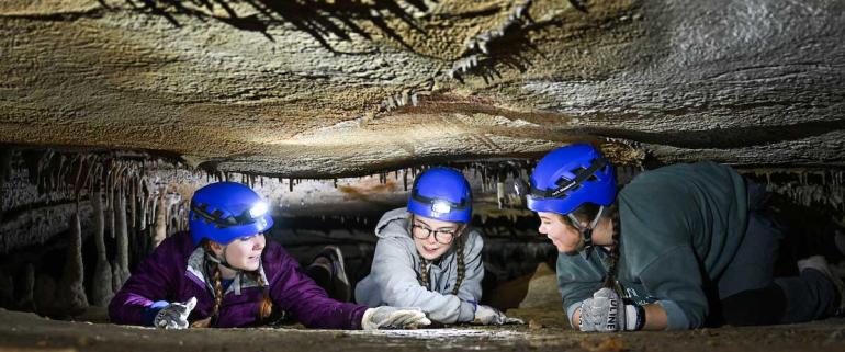 Three UTC students wearing blue hard hats with headlamps crawling in a cave