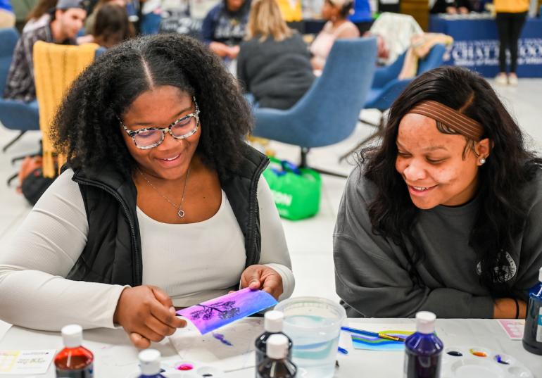Two people work on a colorful art project at a table with paints.