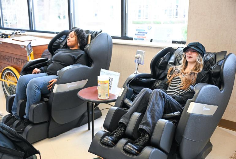 Two people relax in black massage chairs with a small table between them.