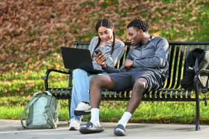 Two students outside on campus Two students, one male and one female, sitting on a bency outside on campus looking at a computer.