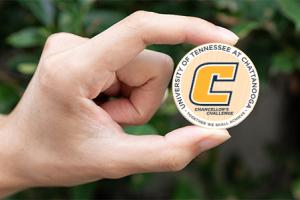 Hand holding a challenge coin that reads "University of Tennessee at Chattanooga. Chancellor's Challenge. Together we shall achieve"