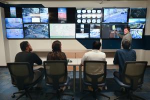 Four people sitting at a table, looking at multiple screens showing transportation data, maps, real-time traffic footage, and more. One person stands and is pointing at one of the screens.