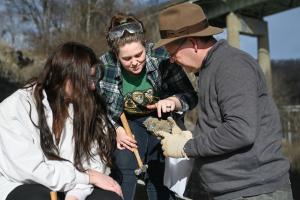Three people outdoors examining a large rock sample together near a bridge. One person wearing safety goggles points at the rock while another holds it with work gloves, and a hammer is visible nearby.