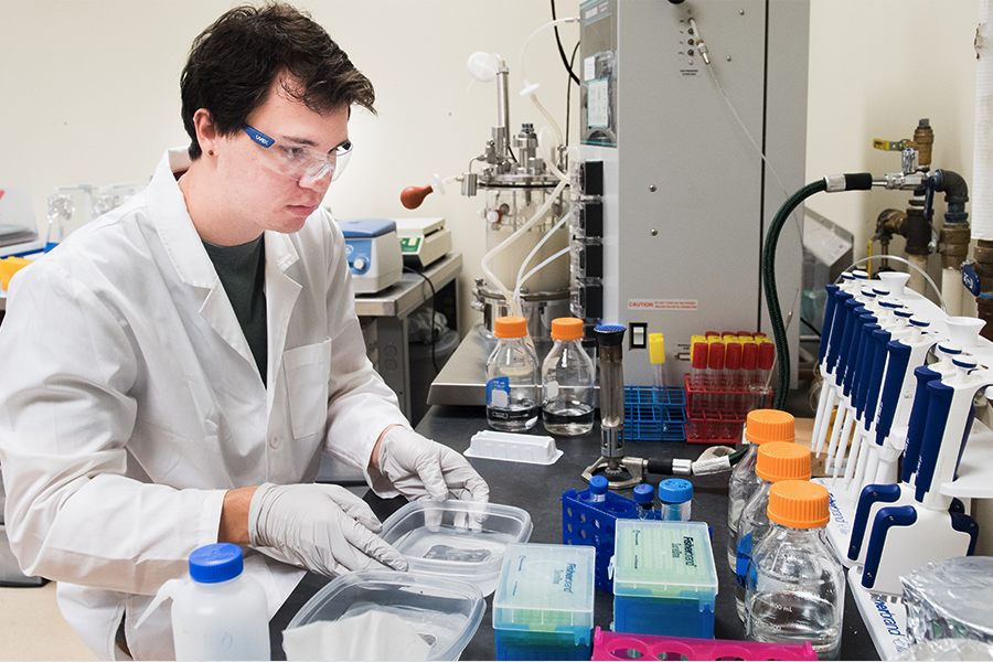 Male chemical engineering student in a white lab coat, goggles, and gloves doing lab work. Male chemical engineering student in a white lab coat, goggles, and gloves doing lab work.