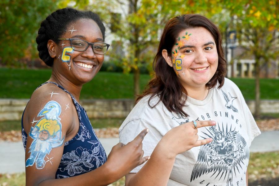 Two female UTC students standing in front of a blurred campus building holding up the Power C hand gesture