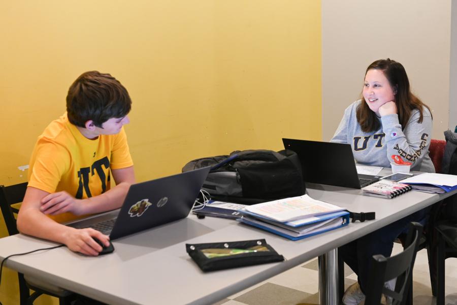 Two UTC students talking at a table with laptops open.