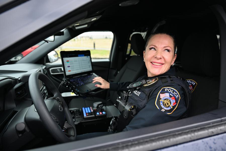 UTC female police officer sitting inside her vehicle looking out the window, her hand is hovering over a laptop 