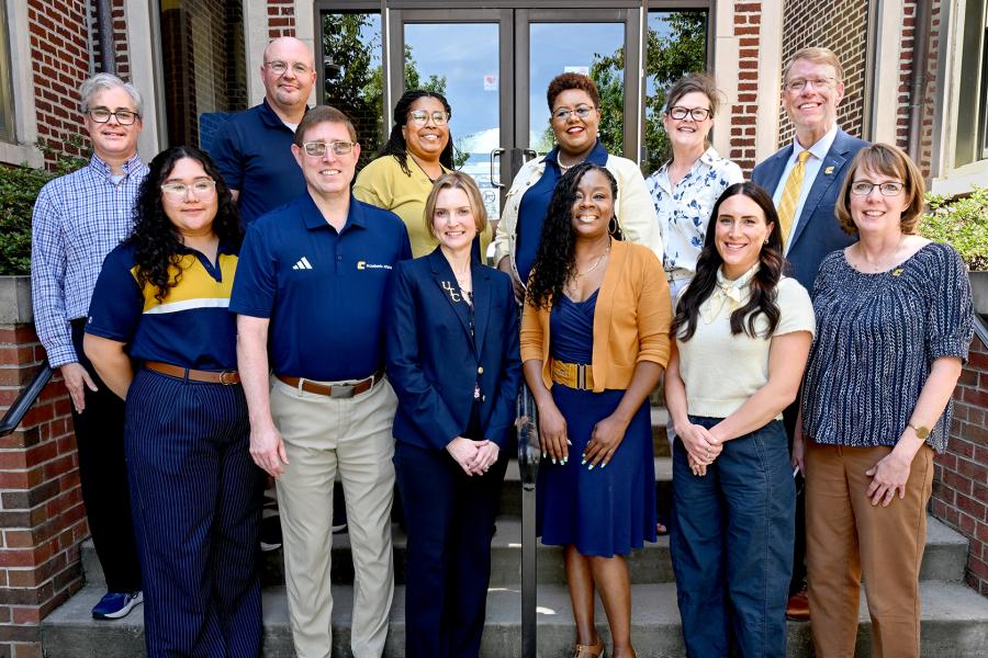 Group portrait of Joel Wells, John Freeze, Delight Yokley, Stephanie Orr, Lauren Ingraham, Matt Matthews, Heidi Maradiaga, Ethan Carver, Elizabeth O'Brien, Charlene Davenport, Rachel Ellis, and Laura Perryman standing on the steps outside Hooper-Race Hall. The group is arranged in two rows in front of the building’s glass entry doors and brick exterior, smiling at the camera in business and business-casual attire.