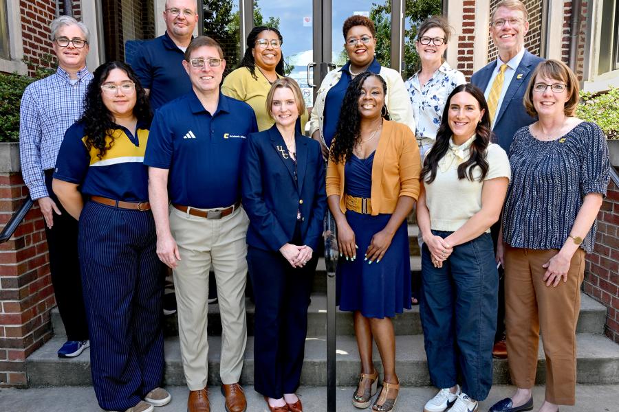 Group portrait of Joel Wells, John Freeze, Delight Yokley, Stephanie Orr, Lauren Ingraham, Matt Matthews, Heidi Maradiaga, Ethan Carver, Elizabeth O'Brien, Charlene Davenport, Rachel Ellis, and Laura Perryman standing on the steps outside Hooper-Race Hall. The group is arranged in two rows in front of the building’s glass entry doors and brick exterior, smiling at the camera in business and business-casual attire.