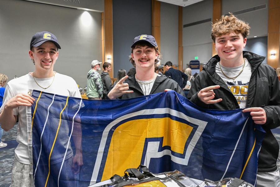 Three UTC male students holding up a blue flag with a gold power C on it while also holding up the power c hand gesture