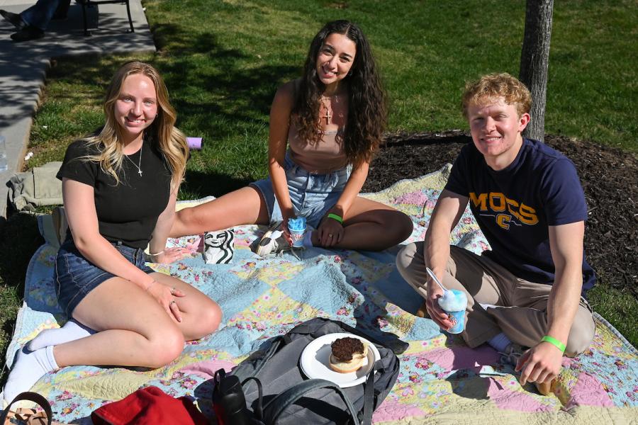 Three UTC students sitting on a blanket outside in the sun eating snacks